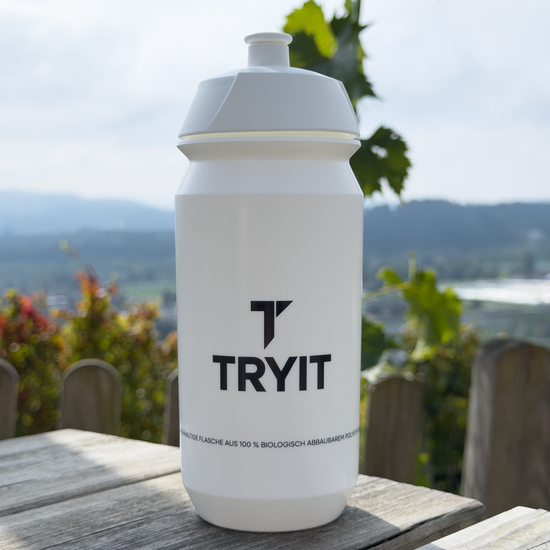 Weiße TRYIT-Sporttrinkflasche auf Holztisch mit verschwommener Landschaft und Weinreben im Hintergrund.