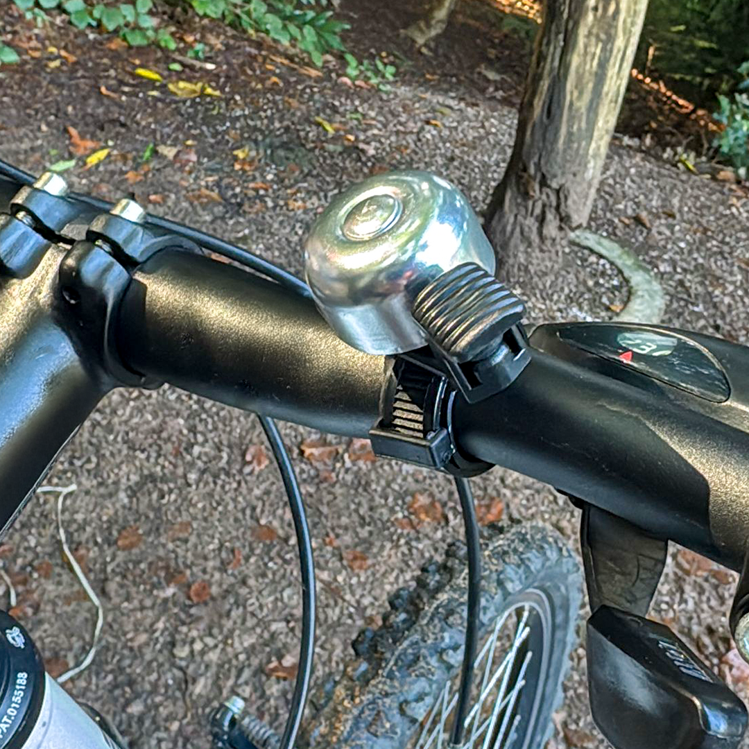 Eine silberne Fahrradklingel ist am schwarzen Lenker eines Mountainbikes befestigt, im Hintergrund Bäume.