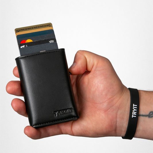 Eine Hand hält eine schwarze Leder-Geldbörse, aus der Kreditkarten herausragen. Am Handgelenk ist ein schwarzes Armband mit der Aufschrift „TRYIT“.