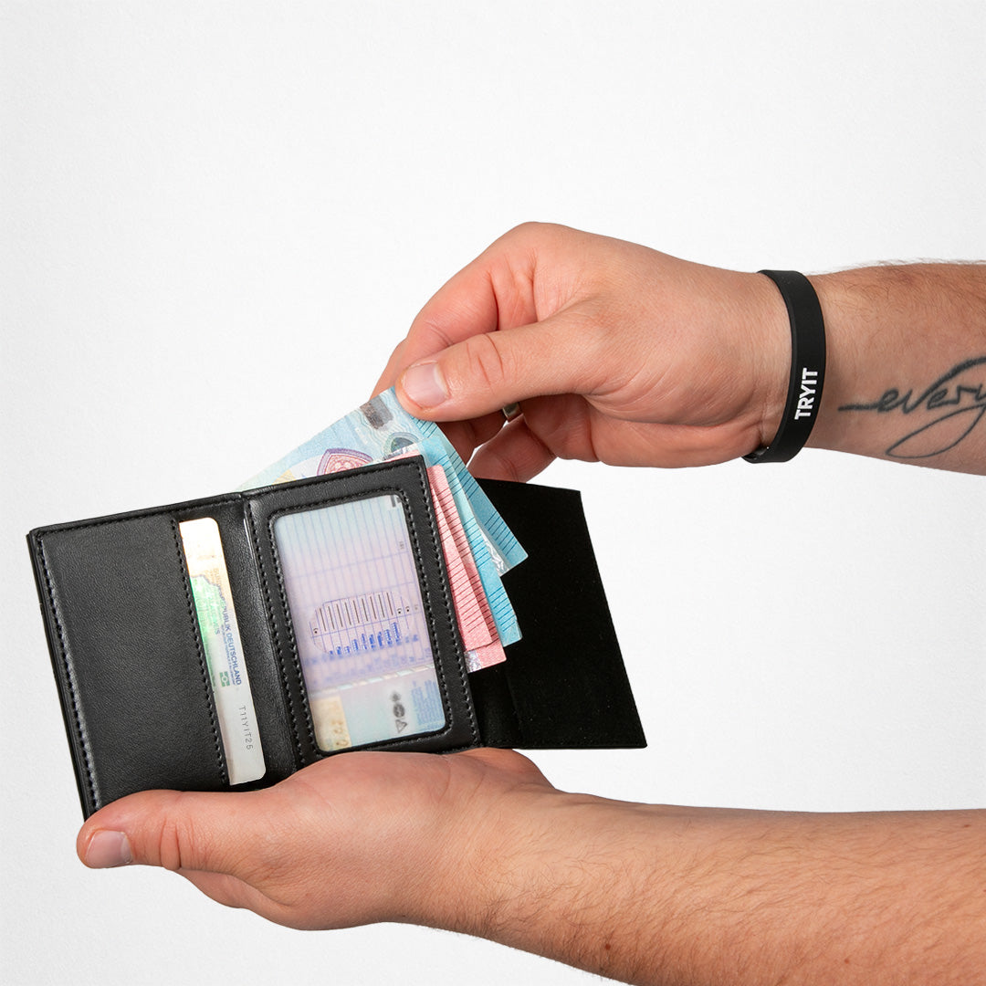 Hände halten eine offene schwarze Ledergeldbörse mit Geldscheinen, Kreditkarten und einem schwarzen Armband mit "TRVTT".