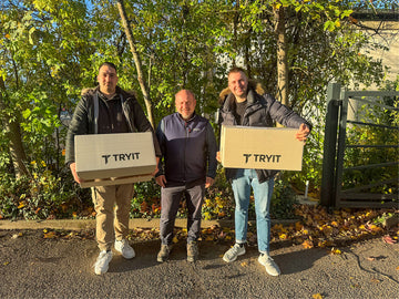 3 Männer in einer Reihe, die beiden Herren links und rechts haben Kartons mit dem Schriftzug "TRYIT" in der Hand und übergeben diese an den Herren in der Mitte
