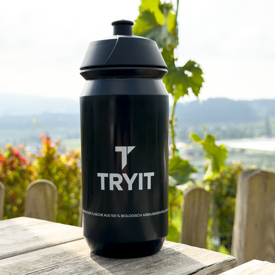 Schwarze Sporttrinkflasche, mit dem weißen "TRYIT"-Logo auf einem Holztisch.